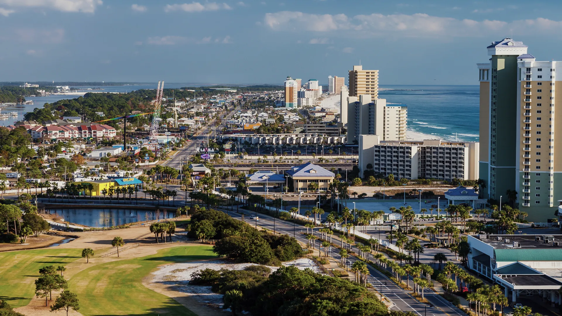 Aerial view of a coastal city with beach, high-rise hotels, roads, golf course, and ocean under a partly cloudy sky.