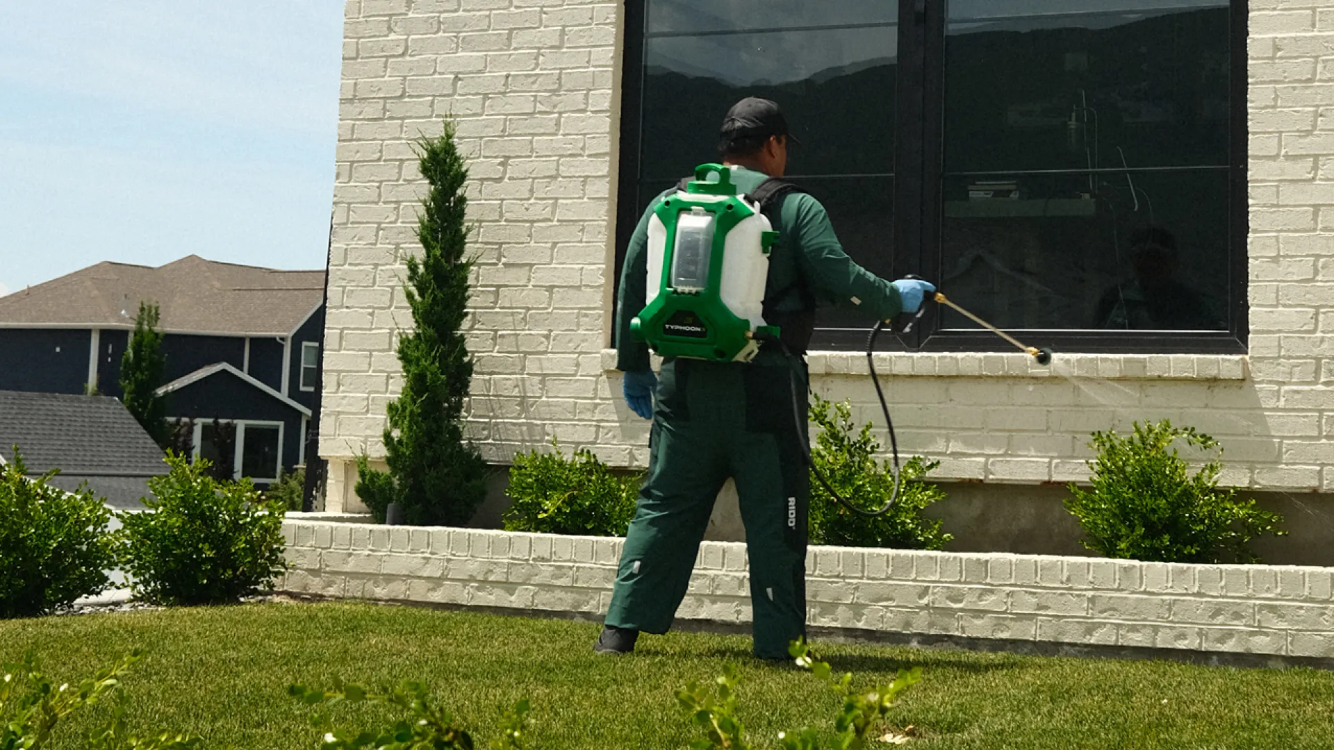 Professional pest control worker spraying insecticide on plants outside a residential home during daytime.