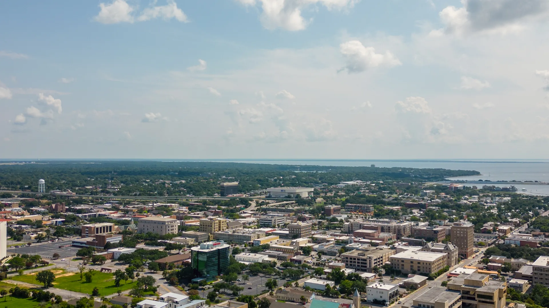Aerial view of a coastal city with buildings, greenery, and a waterfront under a partly cloudy sky.