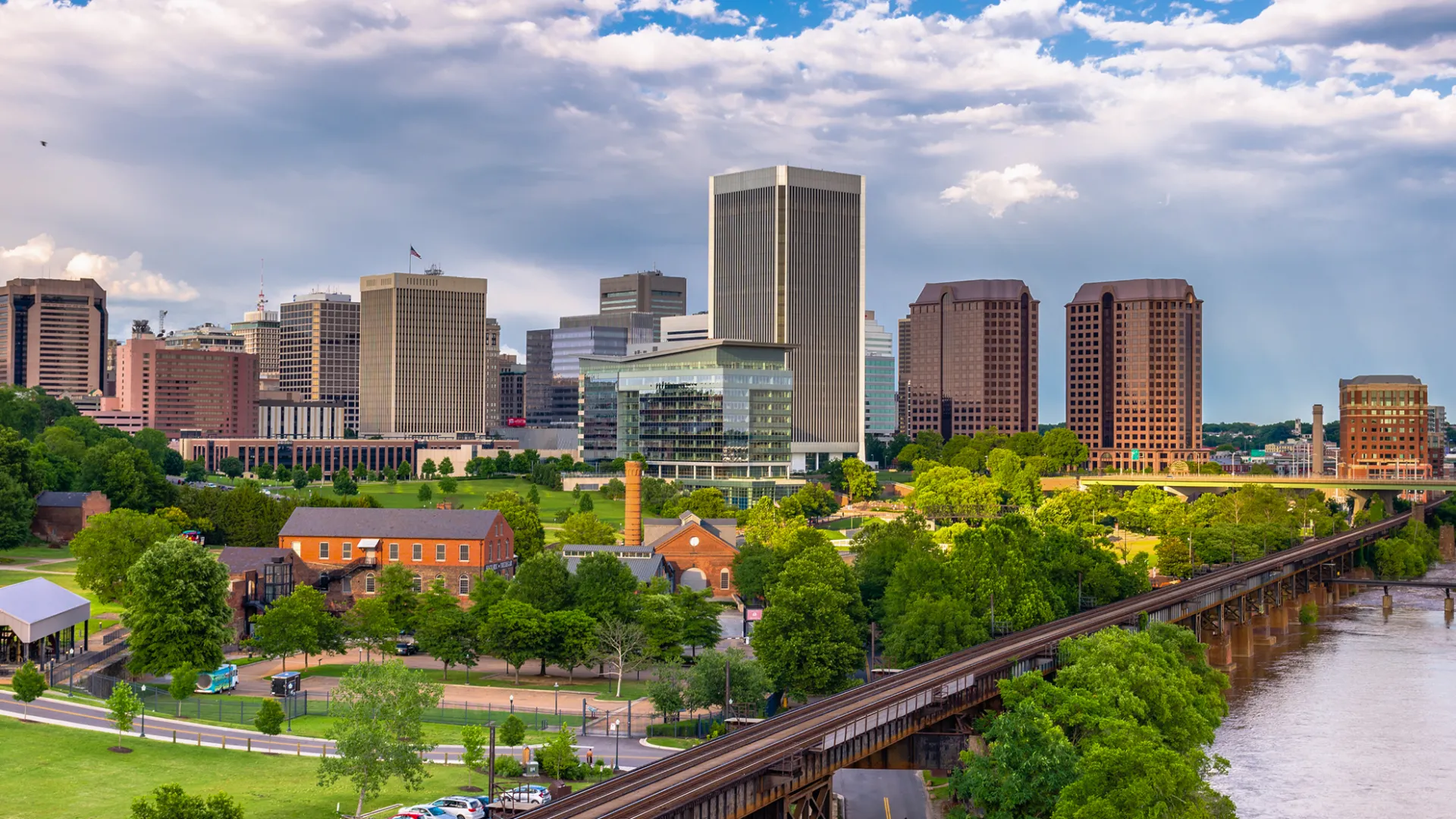 Richmond Virginia skyline with tall buildings, green park, river, and cloudy sky during daytime.