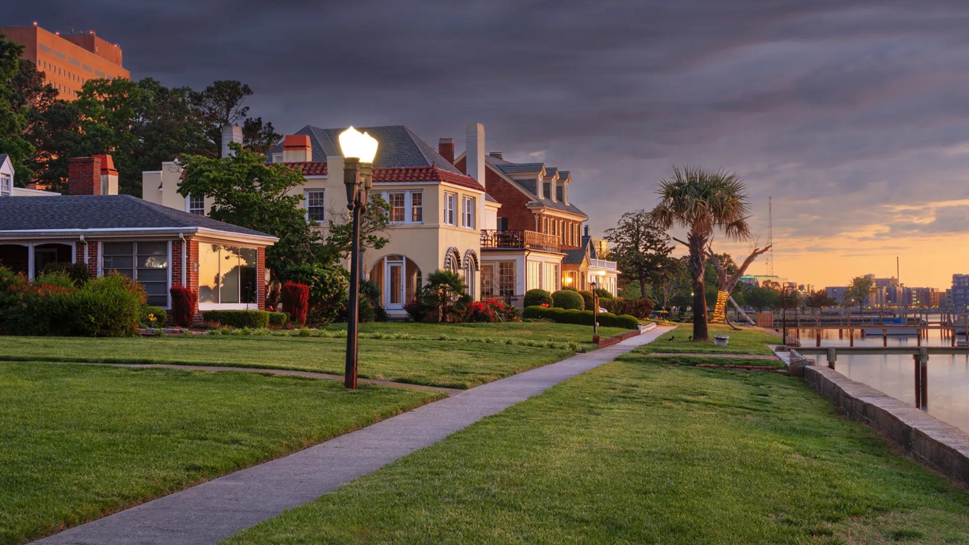 Waterfront street with historic houses, palm trees, and lampposts at sunset under a dramatic cloudy sky.