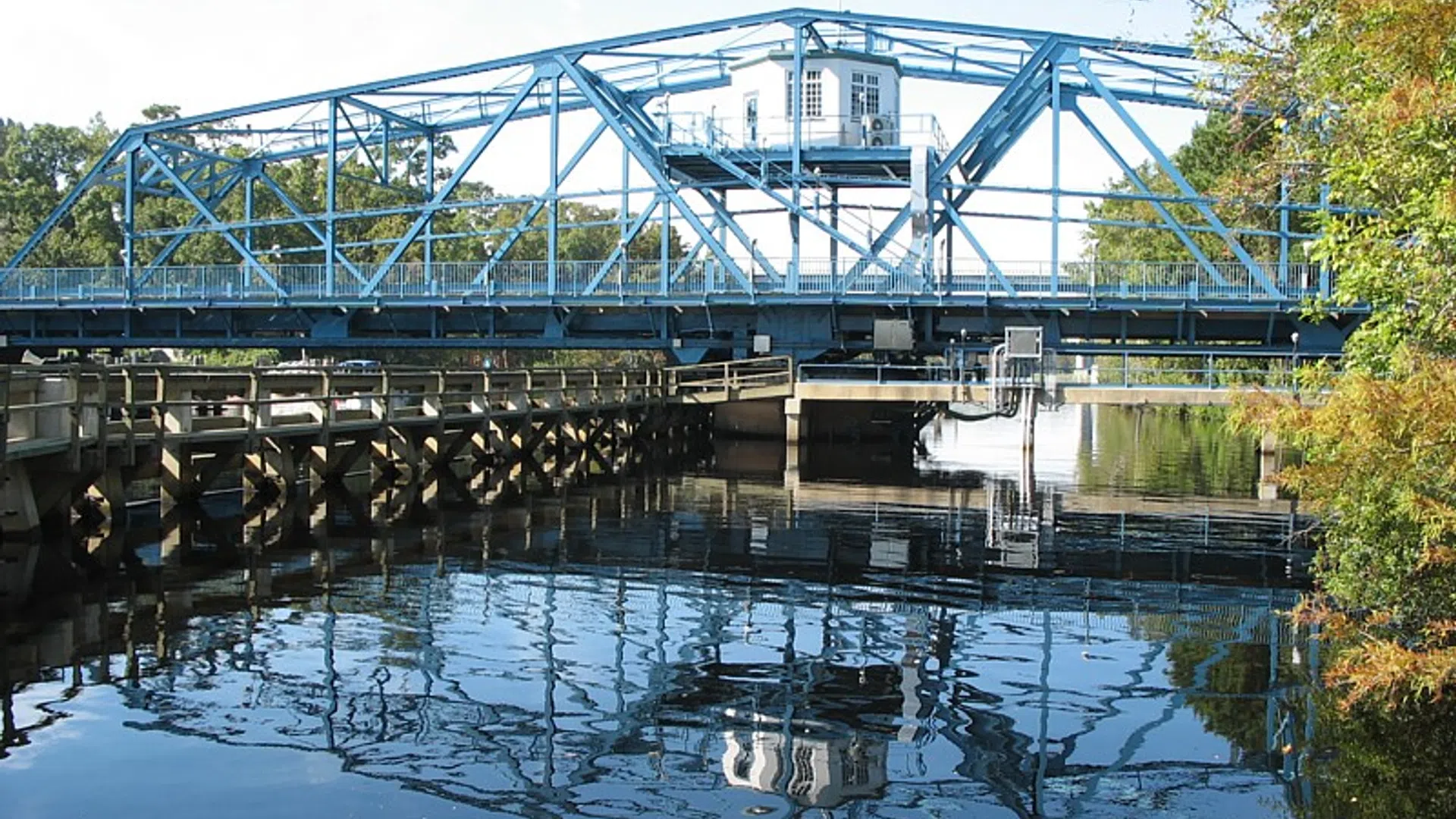 Blue steel drawbridge over calm river water reflecting bridge and surrounding greenery.