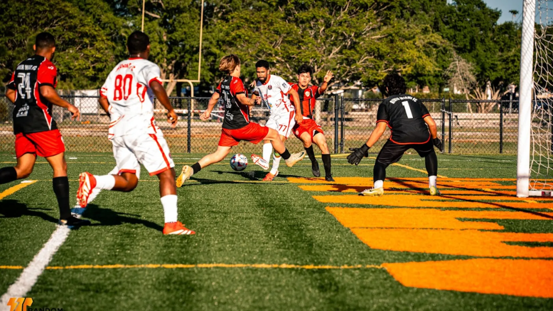 Soccer players in red and white jerseys competing near the goal on a sunny outdoor field with green grass.