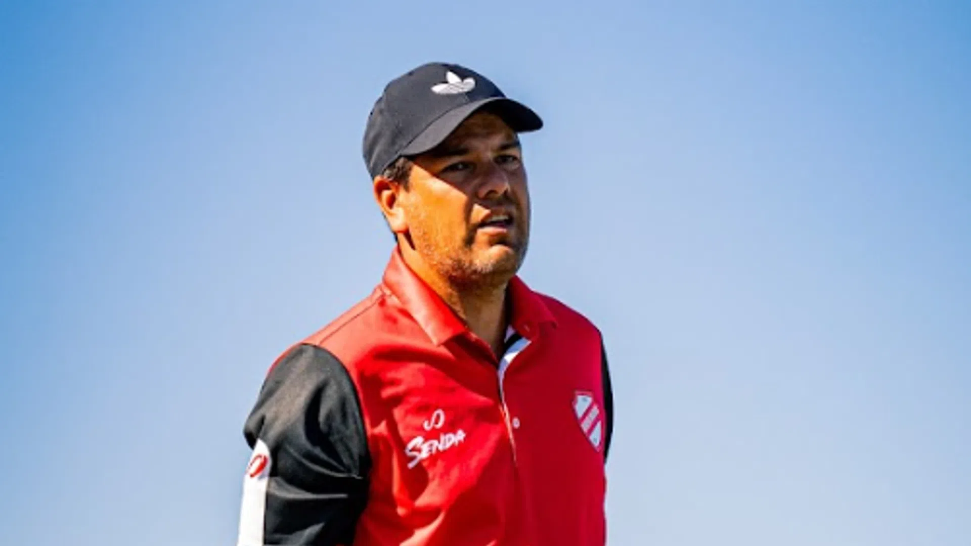 Coach in a red and black sports outfit stands outdoors holding a whistle and a clipboard under clear blue sky.