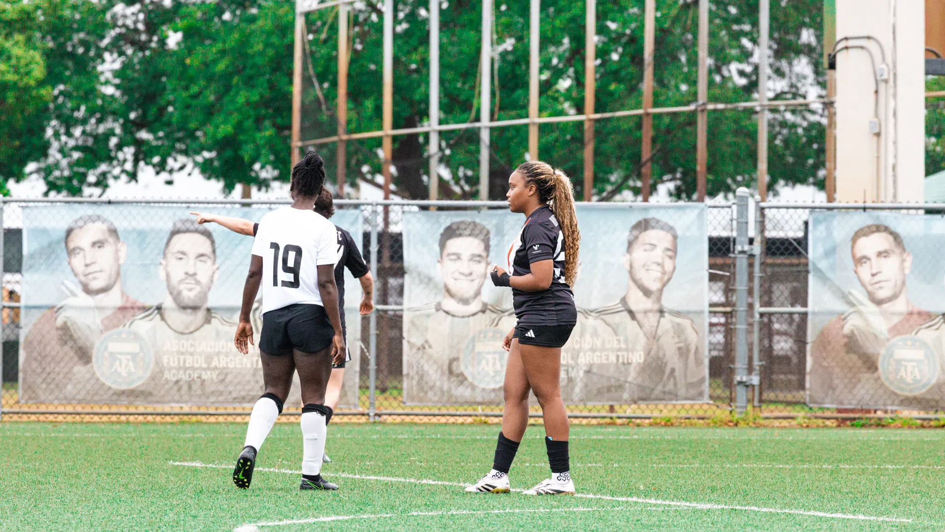 Two female soccer players on field with athletic murals on fence and green trees in background.