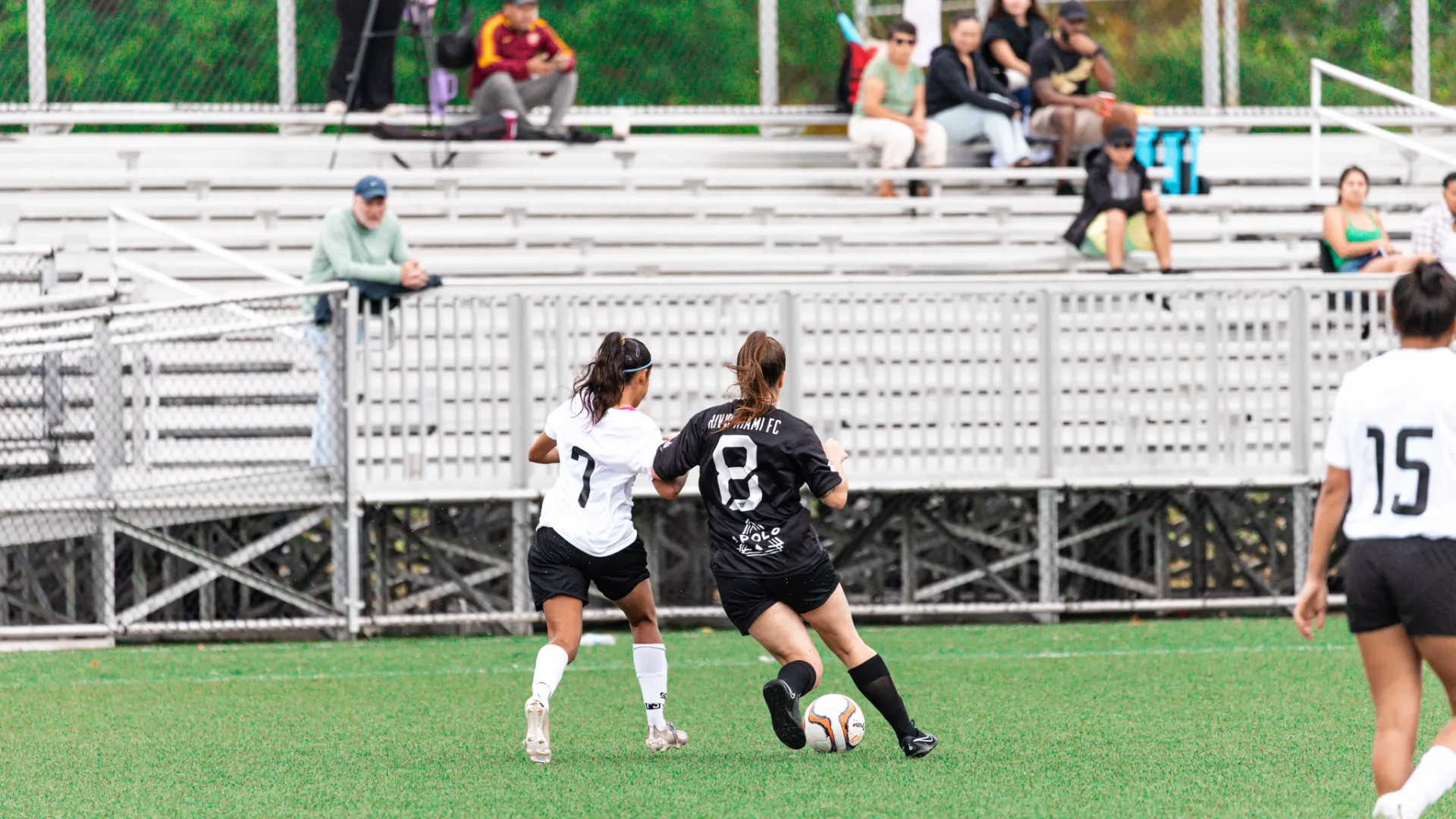 Two female soccer players competing for the ball on a green field with spectators in the background.