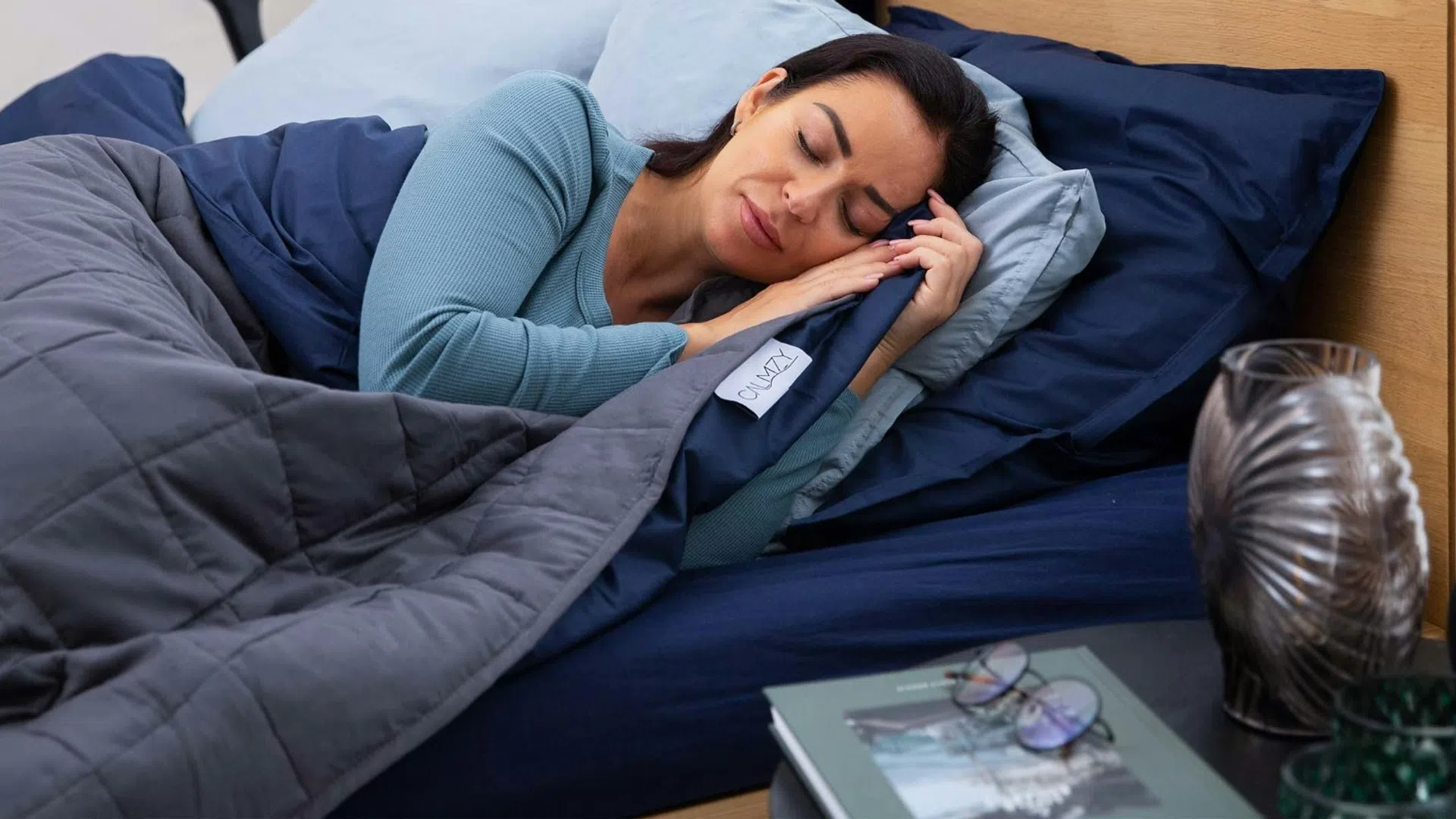 Woman peacefully sleeping in bed with cozy gray and blue bedding in a bright, modern bedroom.