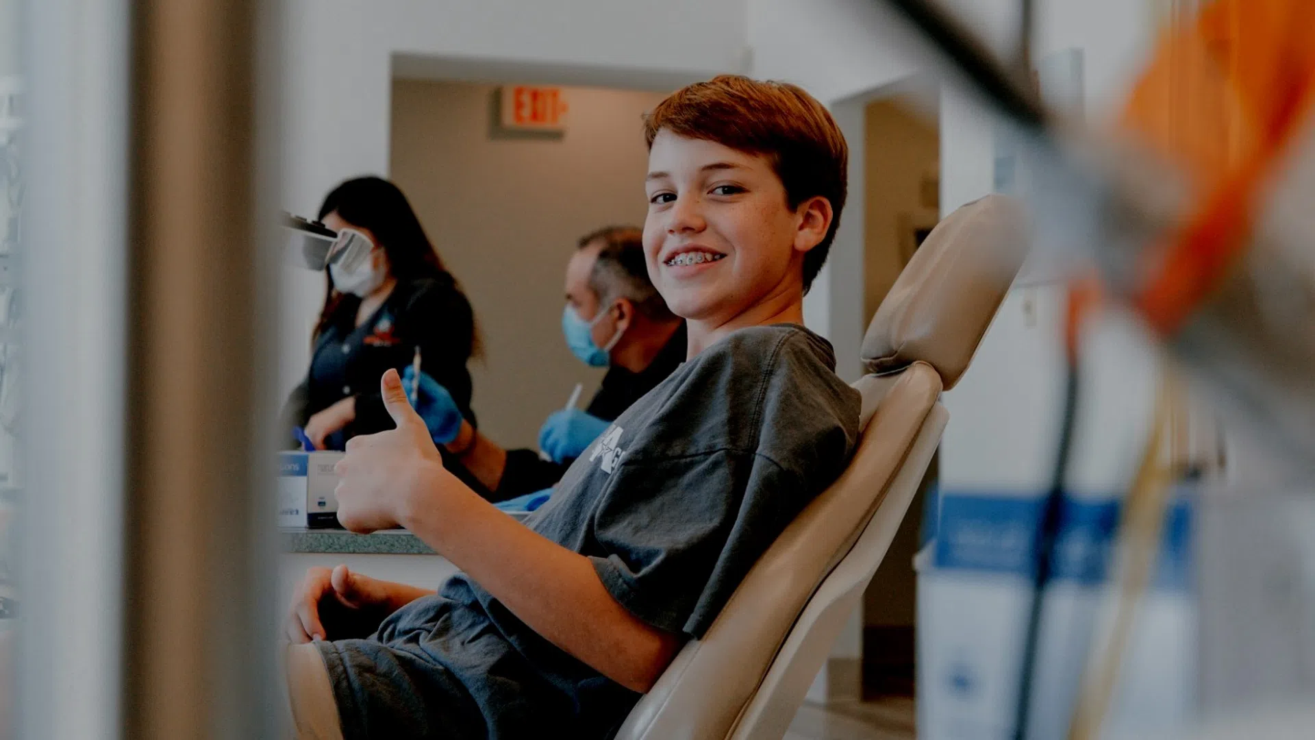 Young boy with braces smiling and giving thumbs up at dental clinic with staff in masks behind him.