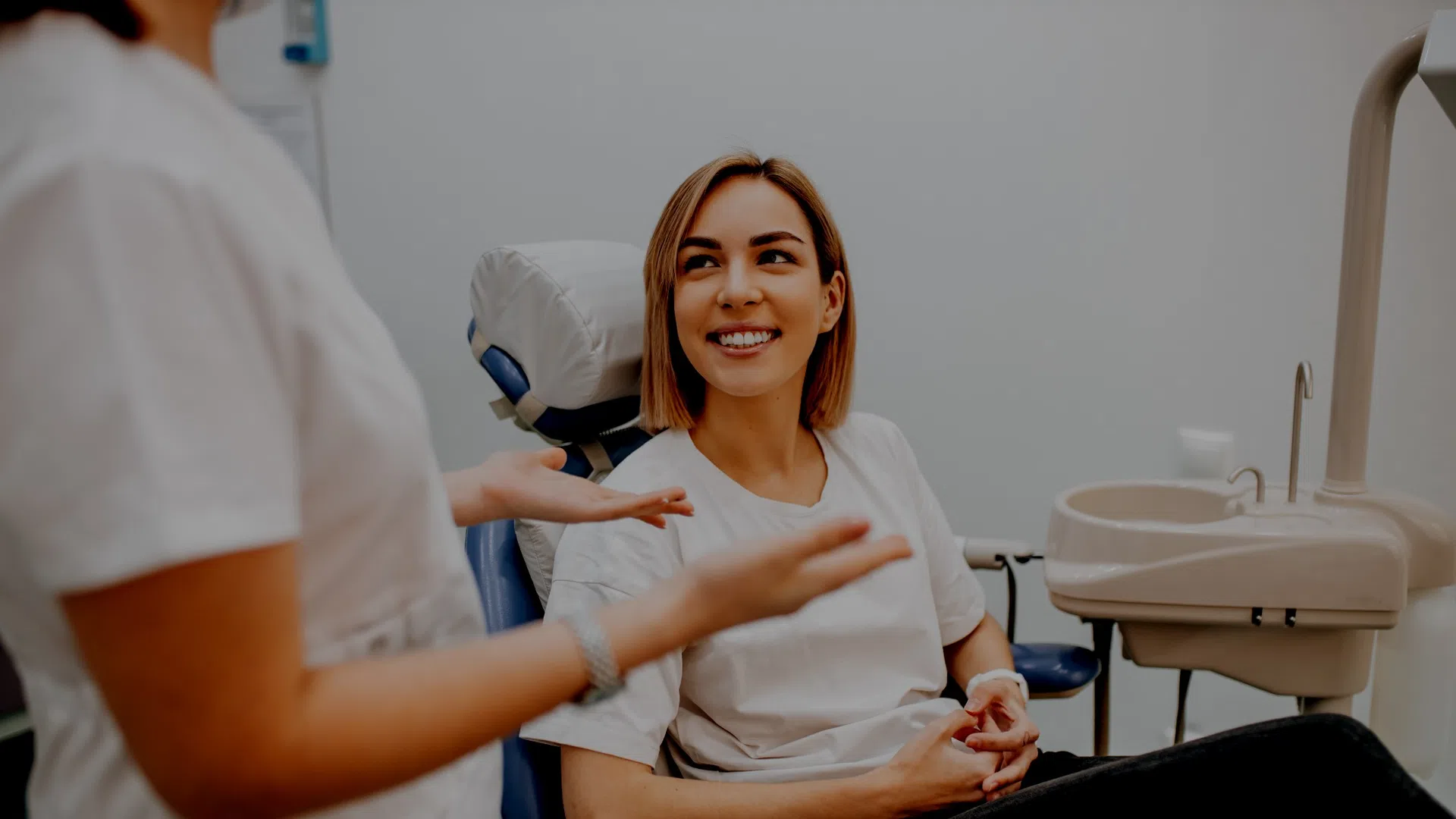 Smiling woman sitting in dental chair talking to a healthcare professional in a bright dental clinic.
