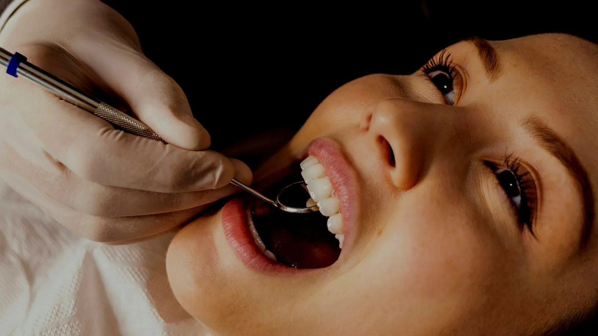 Dentist in gloves examines smiling woman's teeth with dental mirror during checkup appointment.