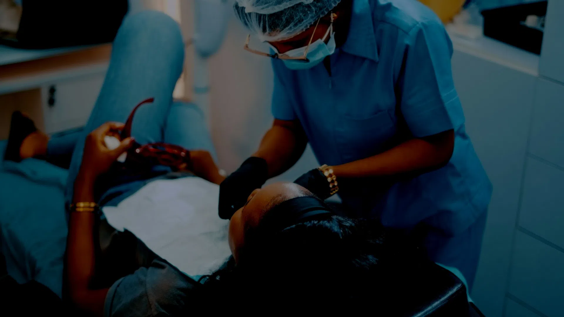 A healthcare professional in blue scrubs and gloves examines a patient lying on a medical chair in a clinic.