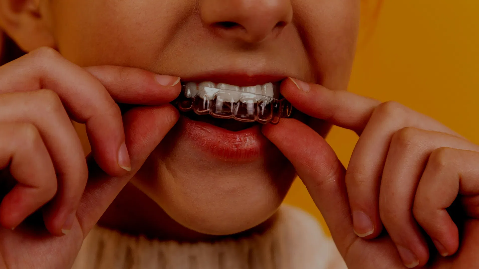 Person placing a clear dental aligner over their teeth against a yellow background.