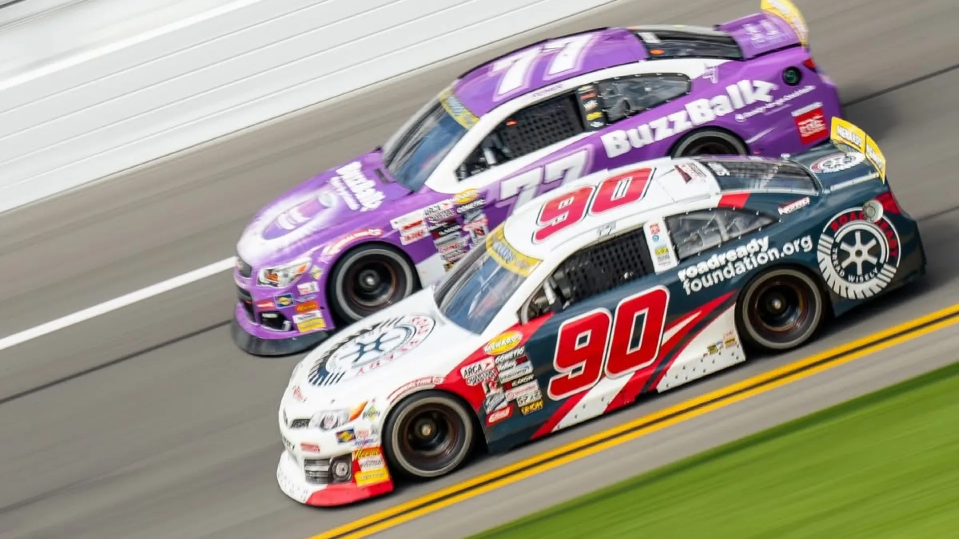 Two stock cars, number 90 and 77, racing side by side on a speedway track with motion blur.