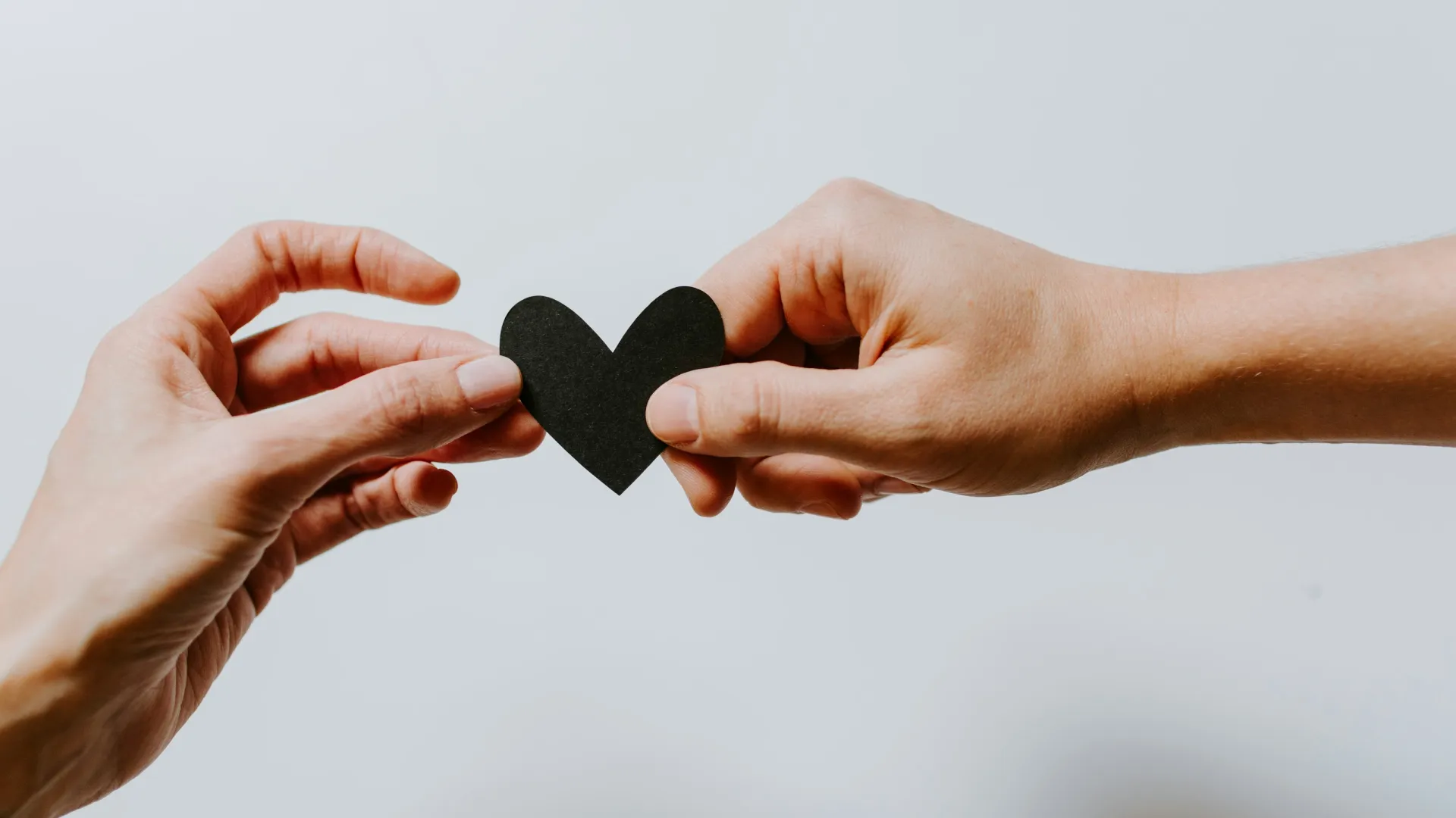 Two hands holding opposite sides of a small black paper heart against a white background.