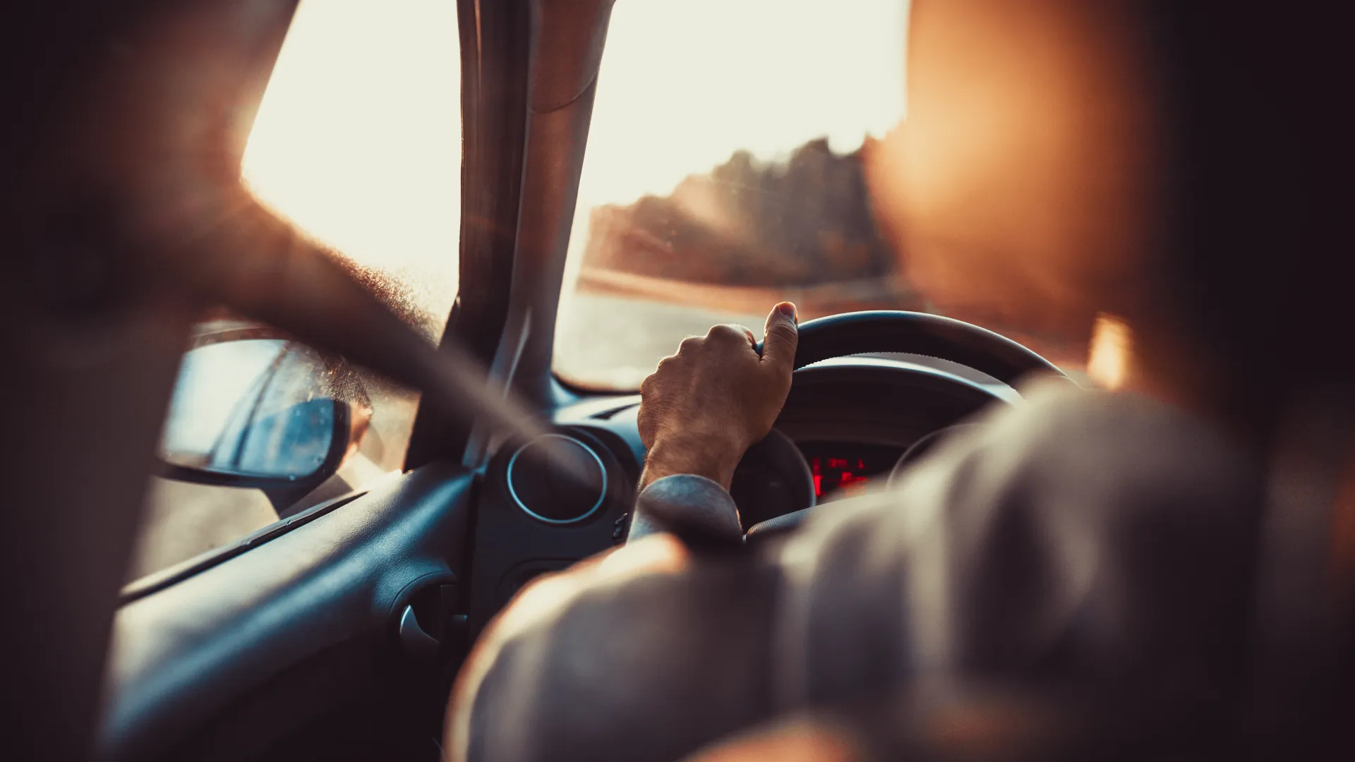 Close-up of a hand gripping a steering wheel, driving into the sunset on a scenic road.