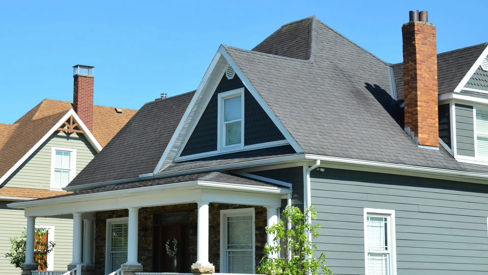 Gray and white house with a front porch, stone pillars, chimneys, and a clear blue sky background