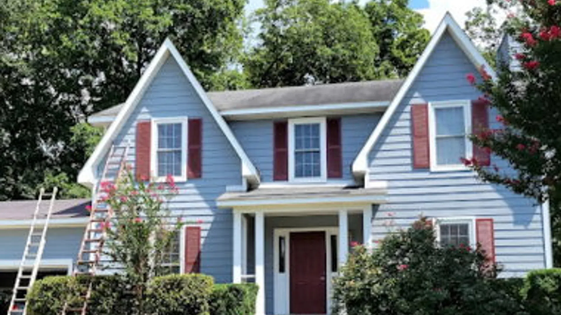 Blue two-story house with white trim and red shutters surrounded by green trees and lawn under a clear sky.