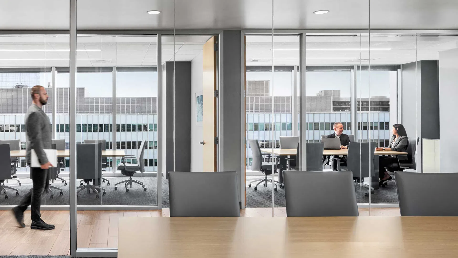 Modern office with glass walls, a man walking, and two colleagues meeting inside a conference room.