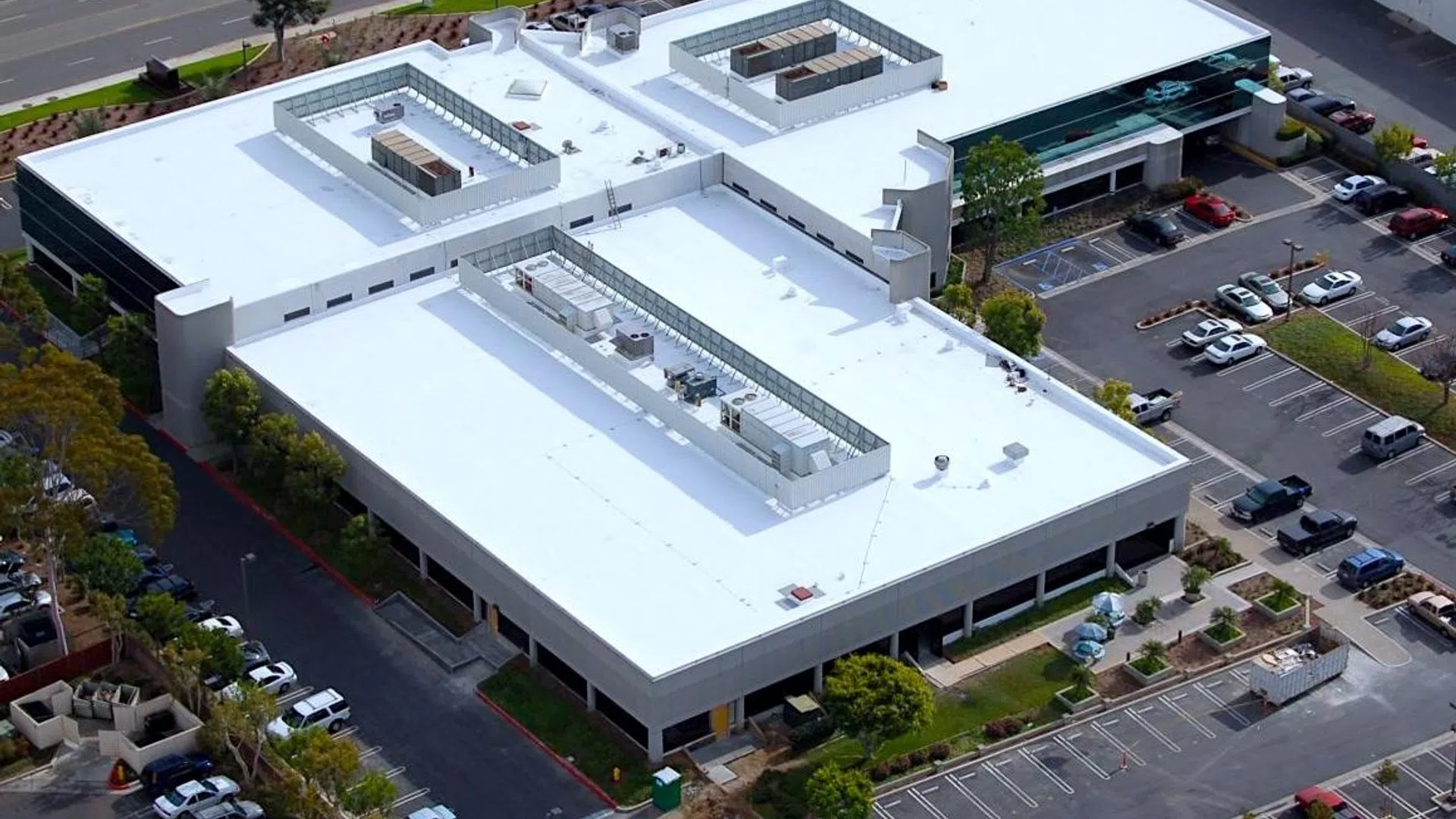 Aerial view of a large commercial building with flat white roofing and surrounding parking lots and greenery.