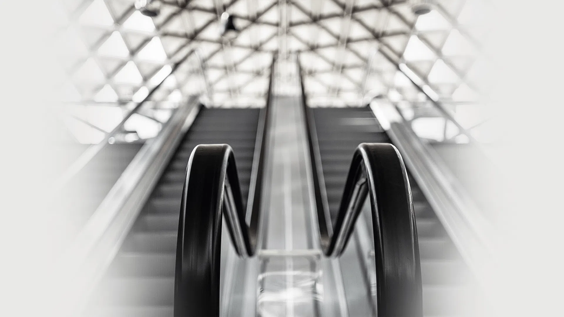 Black handrails and steps of a moving escalator with a bright geometric ceiling pattern above.