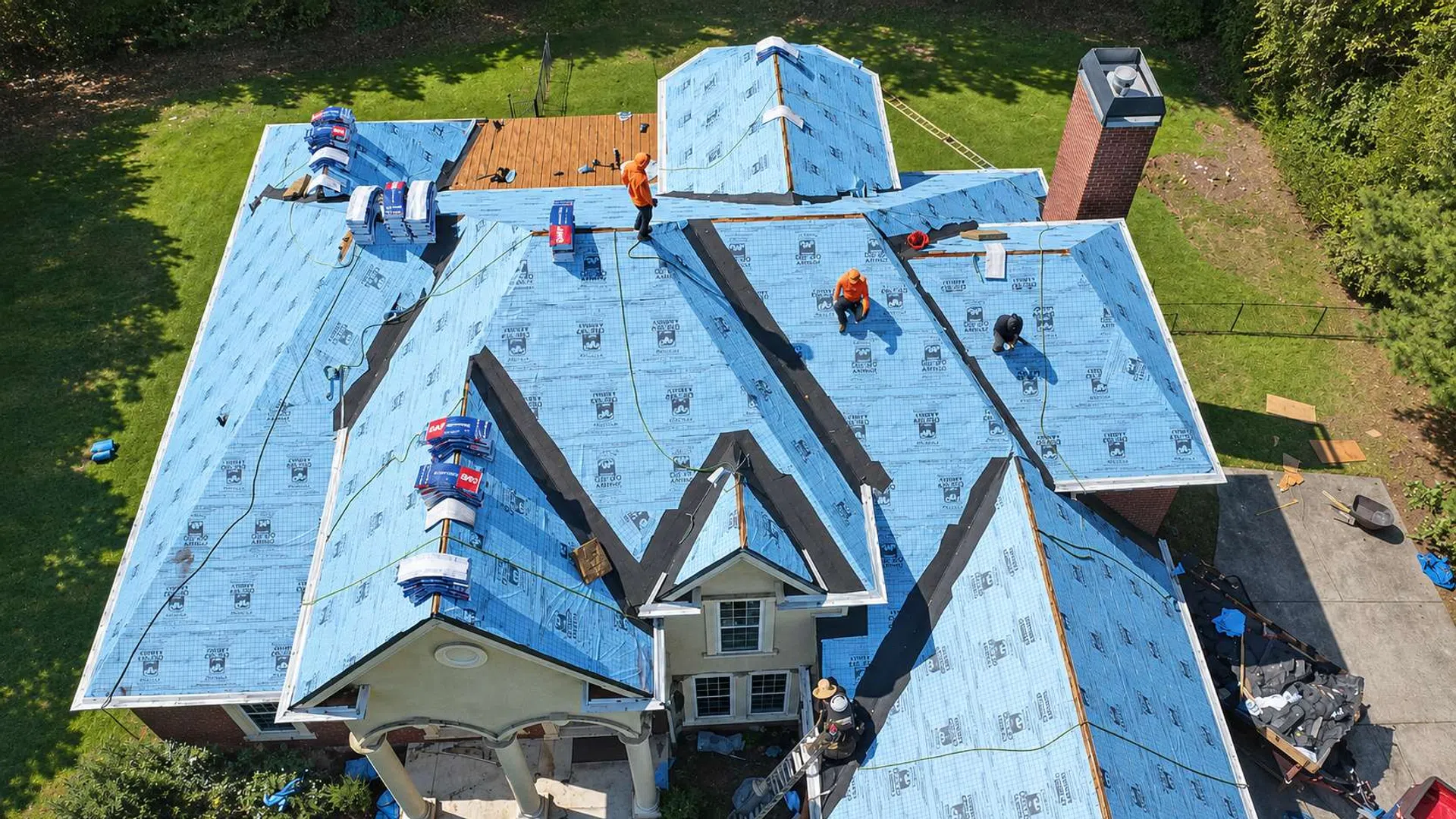 Aerial view of workers installing blue underlayment on a large residential roof under clear weather conditions.