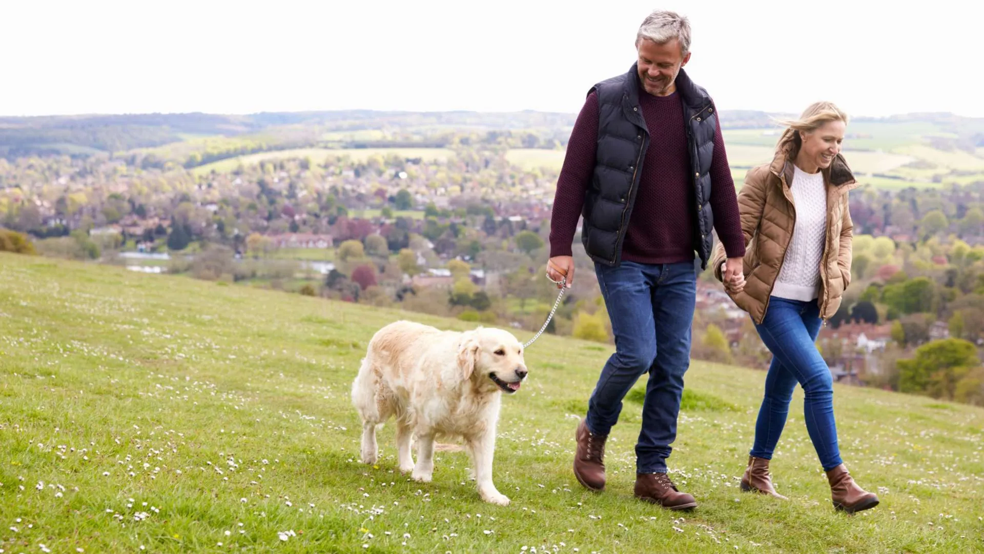 Happy mature couple walking a golden retriever dog on a hill with a scenic village view in the background