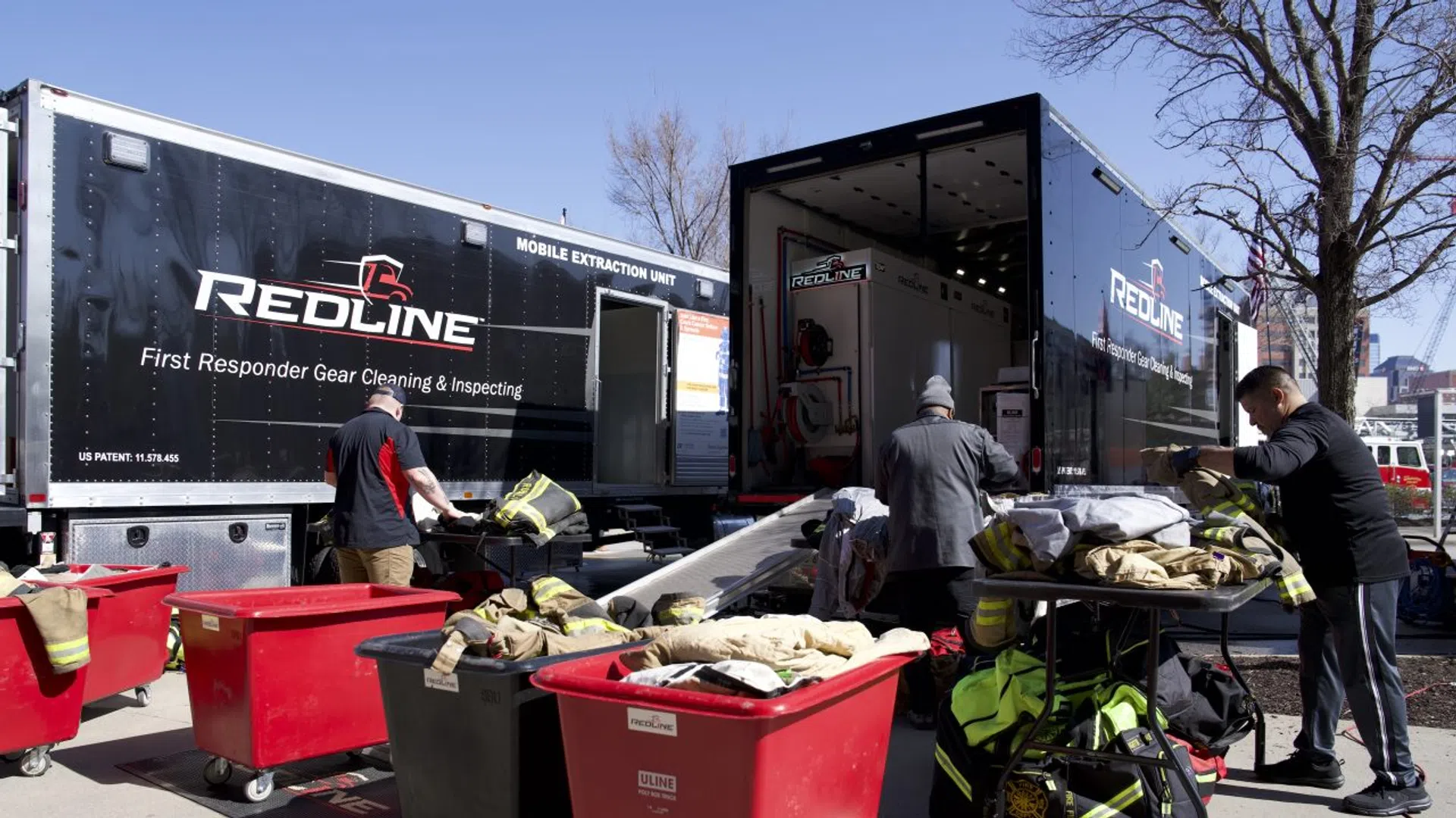 Workers inspecting and cleaning first responder gear using Redline mobile extraction units outdoors on a sunny day.