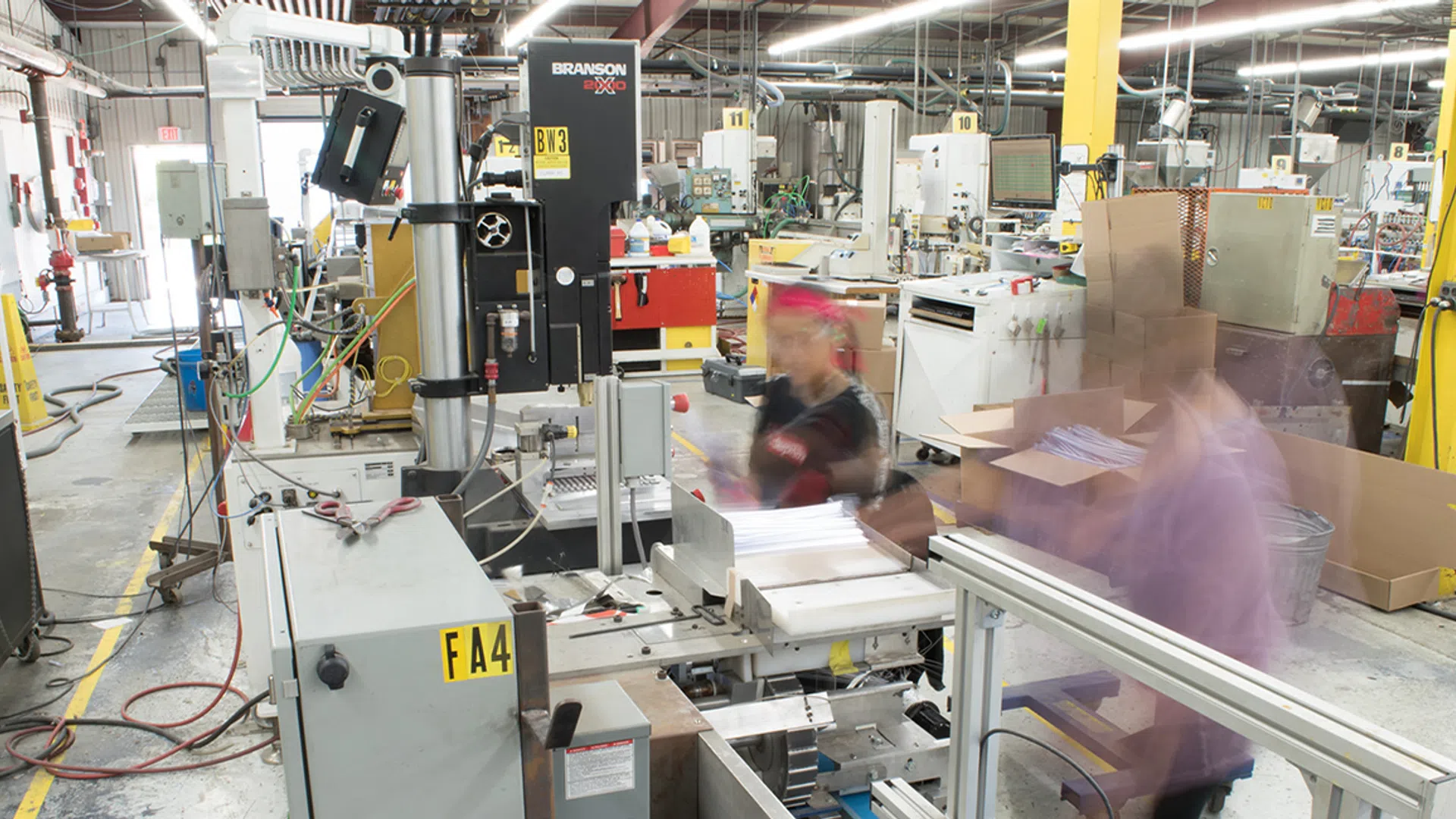 Busy factory floor with machinery and workers assembling products amid industrial equipment and stacked boxes