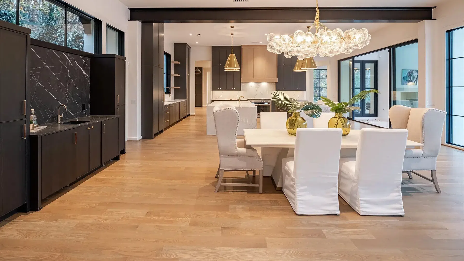 Modern open-concept dining and kitchen area with white chairs, wooden floors, and large glass windows.