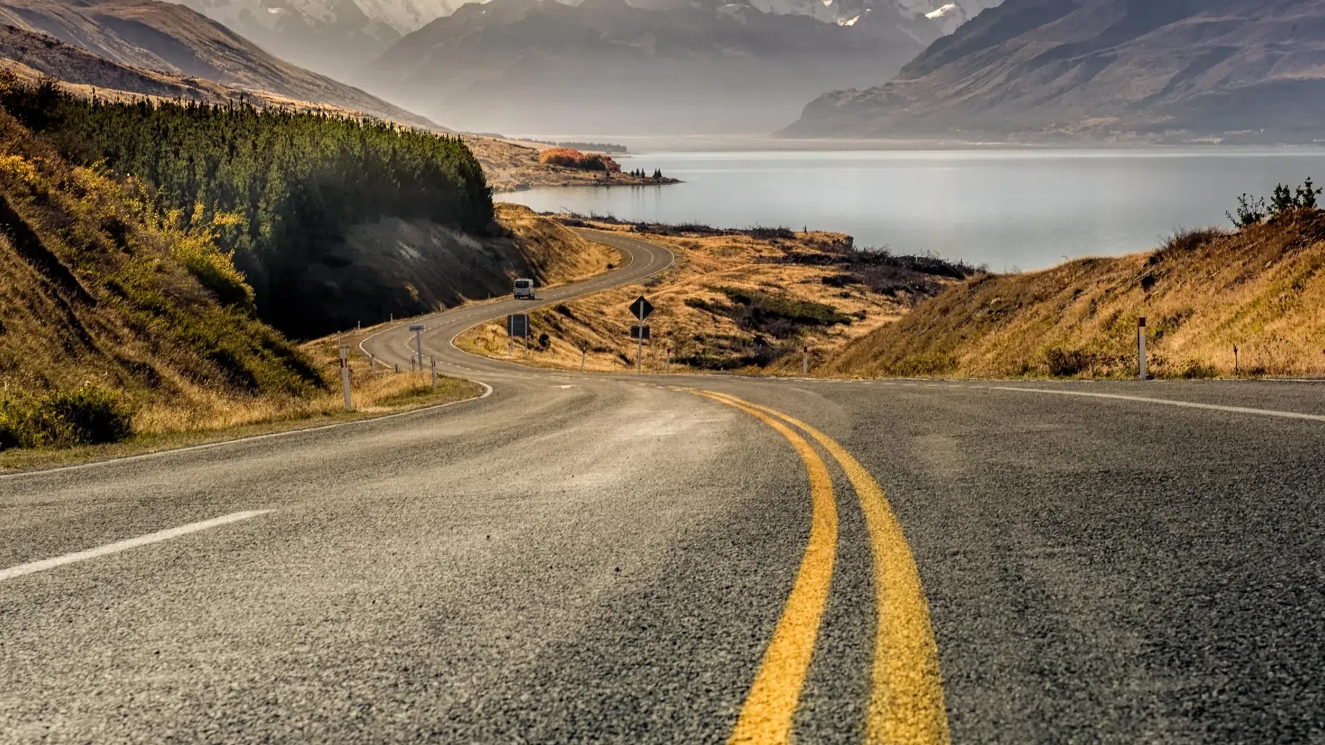Open winding road beside lake with mountains in the distance under cloudy sky during autumn.