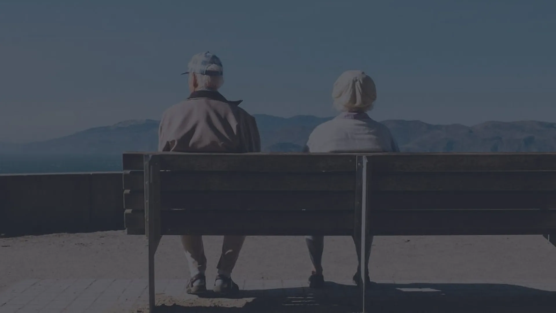 Elderly couple sitting on a bench facing mountains under clear blue sky, enjoying peaceful outdoor view.