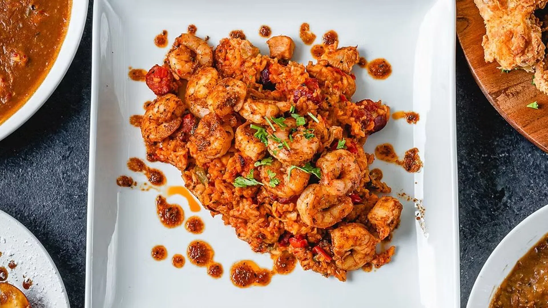 Variety of southern dishes including shrimp and rice, fried chicken, red beans and rice, and dessert on dark table.