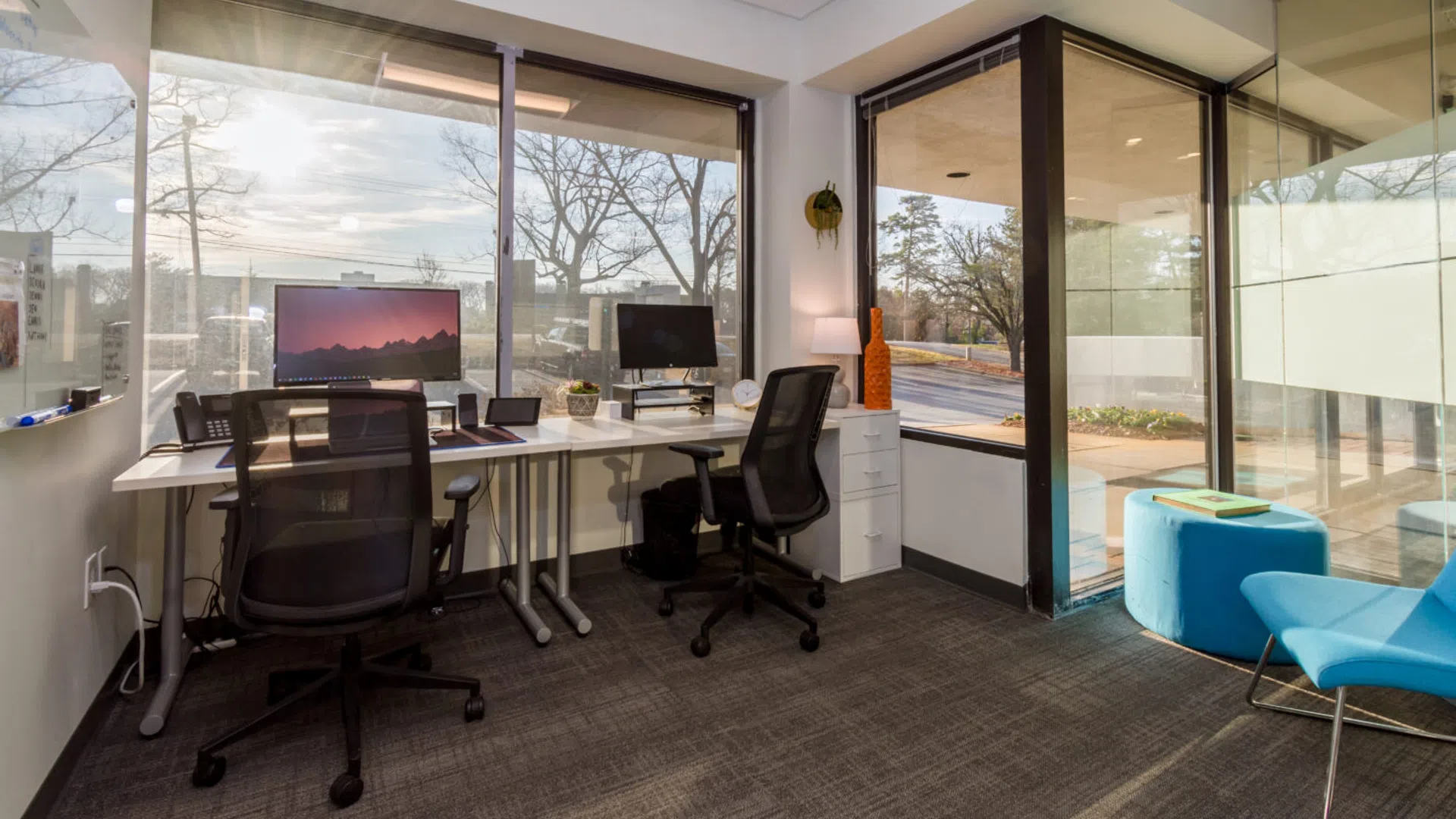 Modern office with dual monitors, ergonomic chairs, large windows, and natural light overlooking trees and street.