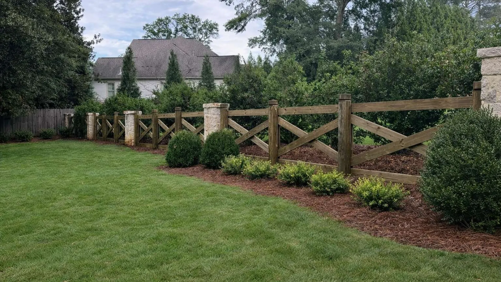 Wooden cross fence with stone pillars borders a landscaped garden with green grass and shrubs under a cloudy sky