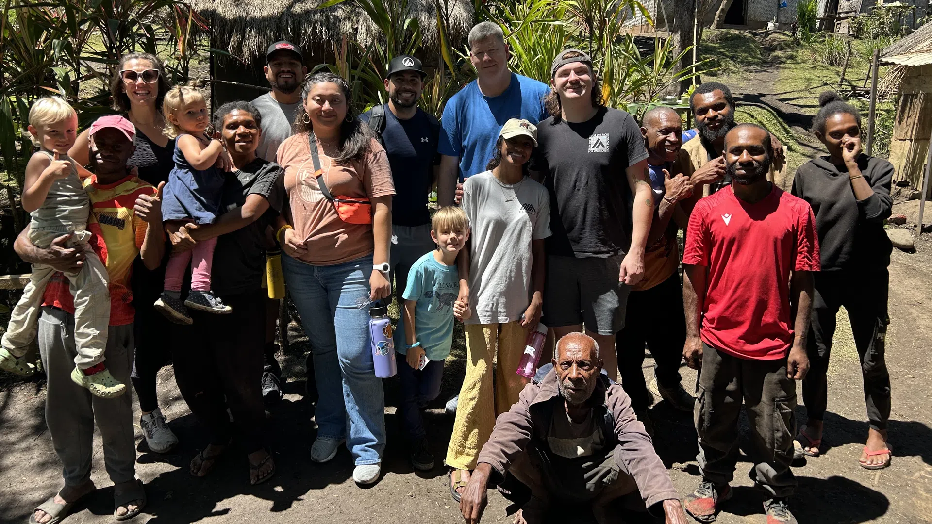 Diverse group of adults and children standing outdoors in front of trees and rural huts on a sunny day.
