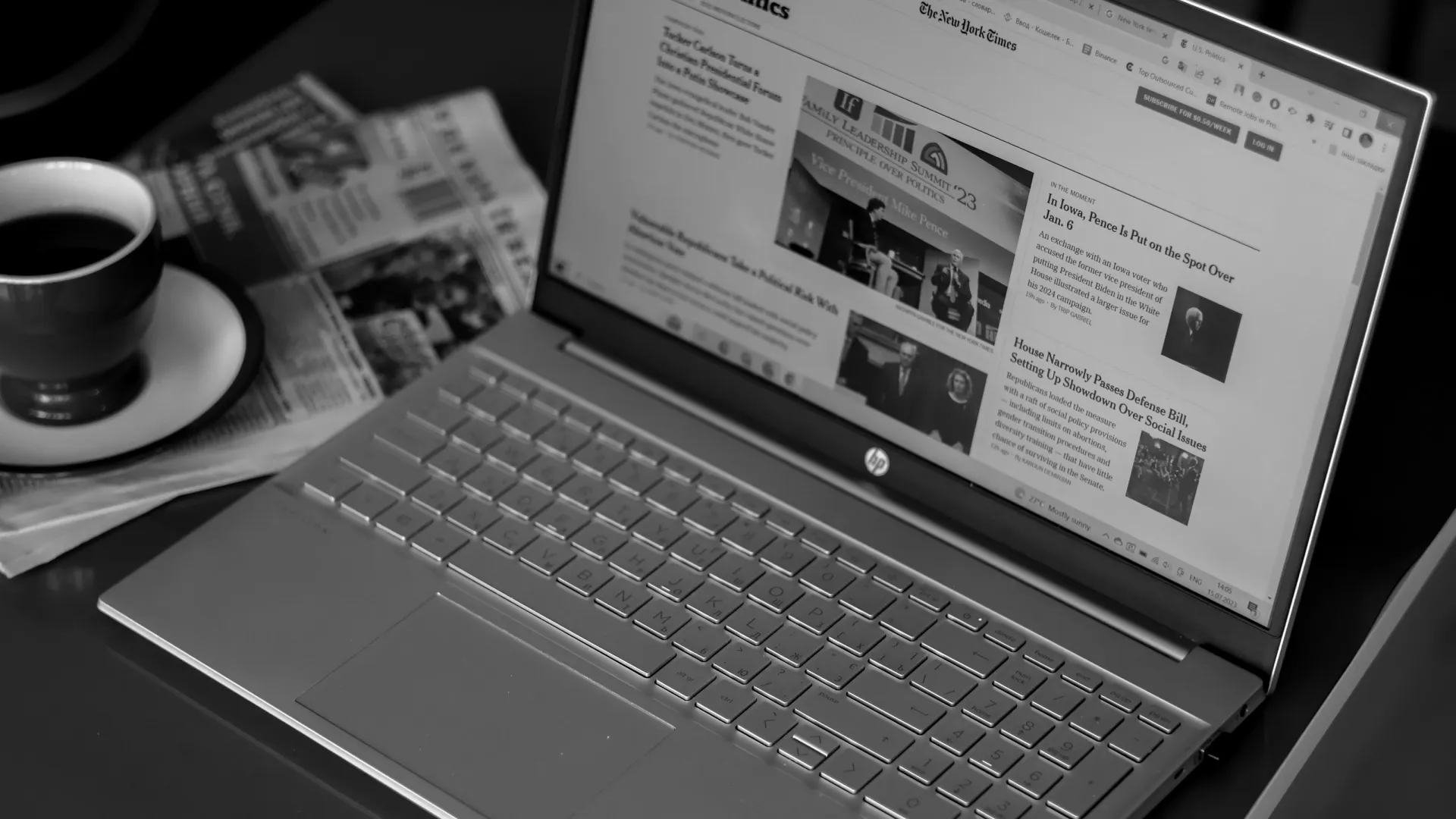 Black and white image of a laptop displaying a news website with a coffee cup and newspapers nearby.