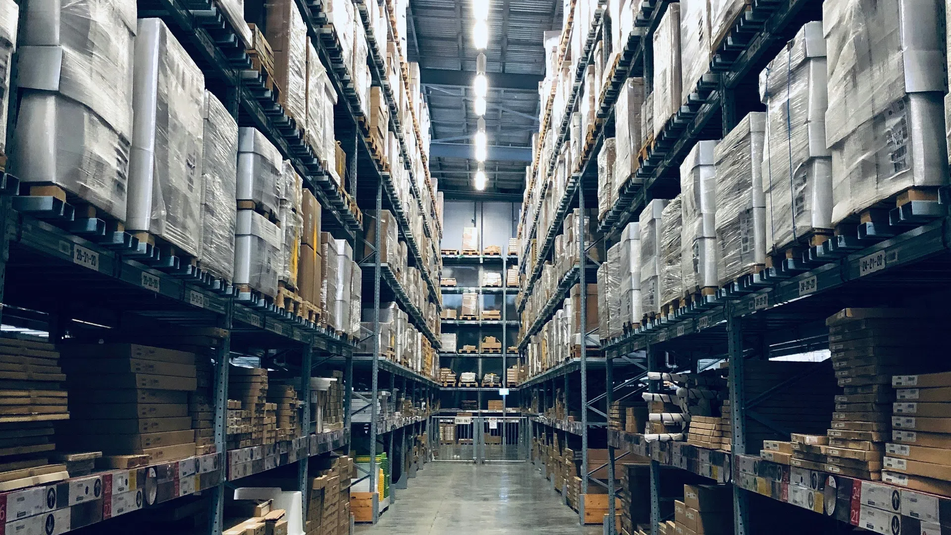 Long aisle view of a large warehouse with tall shelves stacked with pallets and boxes under bright ceiling lights