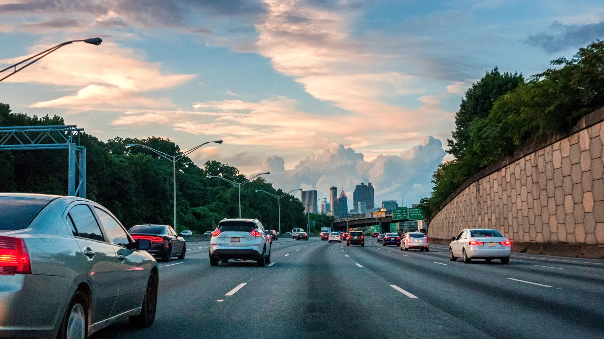 Busy multi-lane highway with cars driving towards a city skyline under a colorful sunset sky.