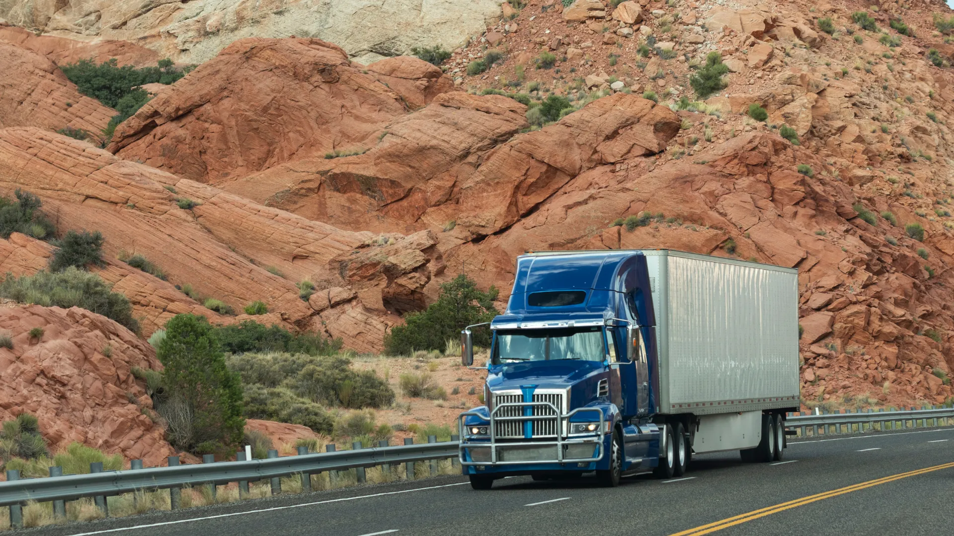 Blue semi-truck driving on highway through red rock desert landscape with sparse vegetation.