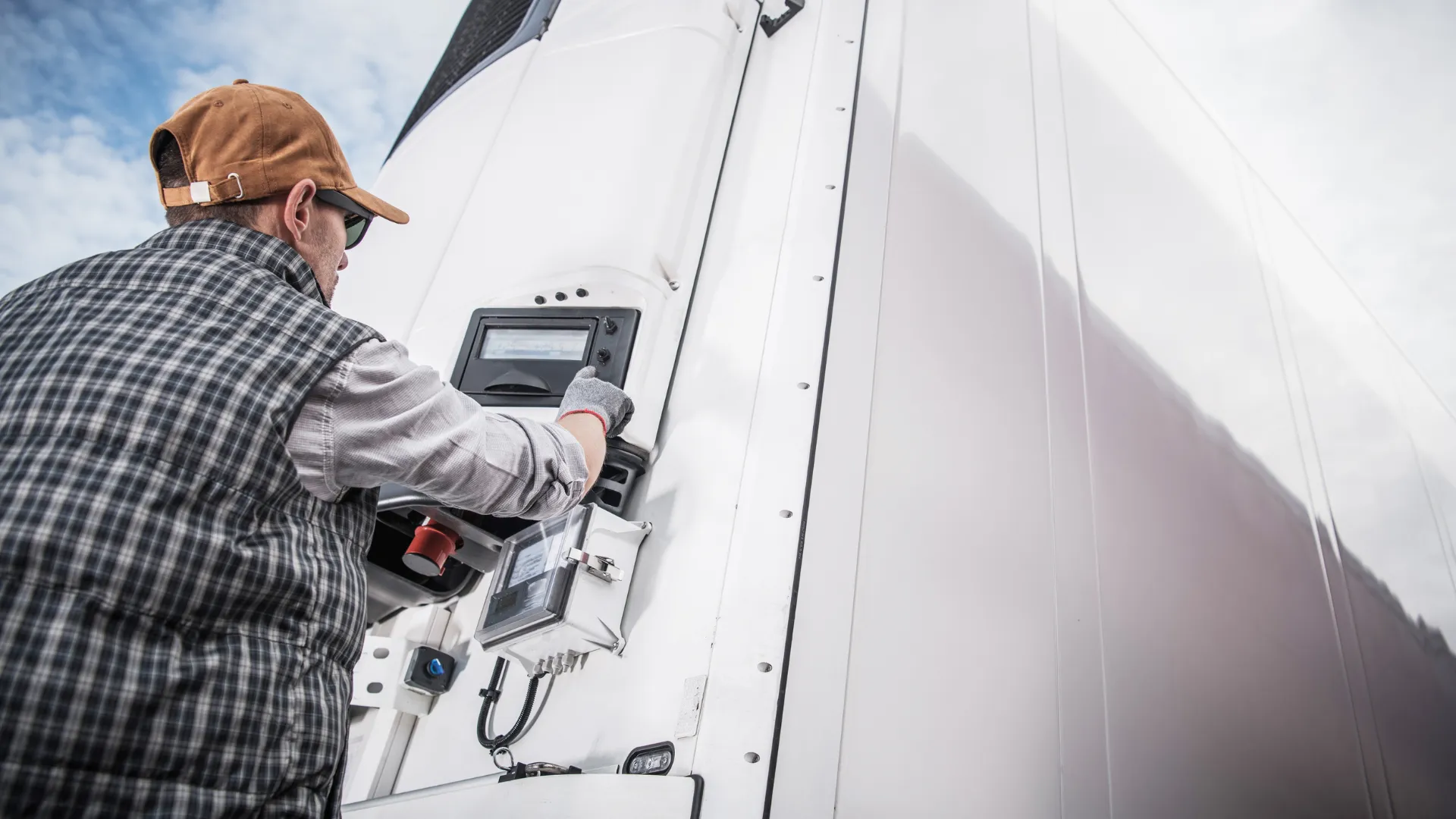 Man in plaid shirt and cap adjusting control panel on large white refrigerated truck under cloudy sky