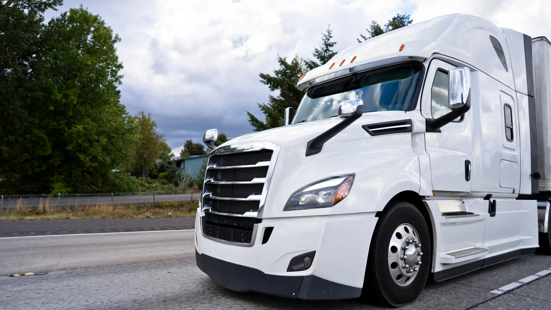 White semi-truck driving on highway with cloudy sky and green trees along the roadside.