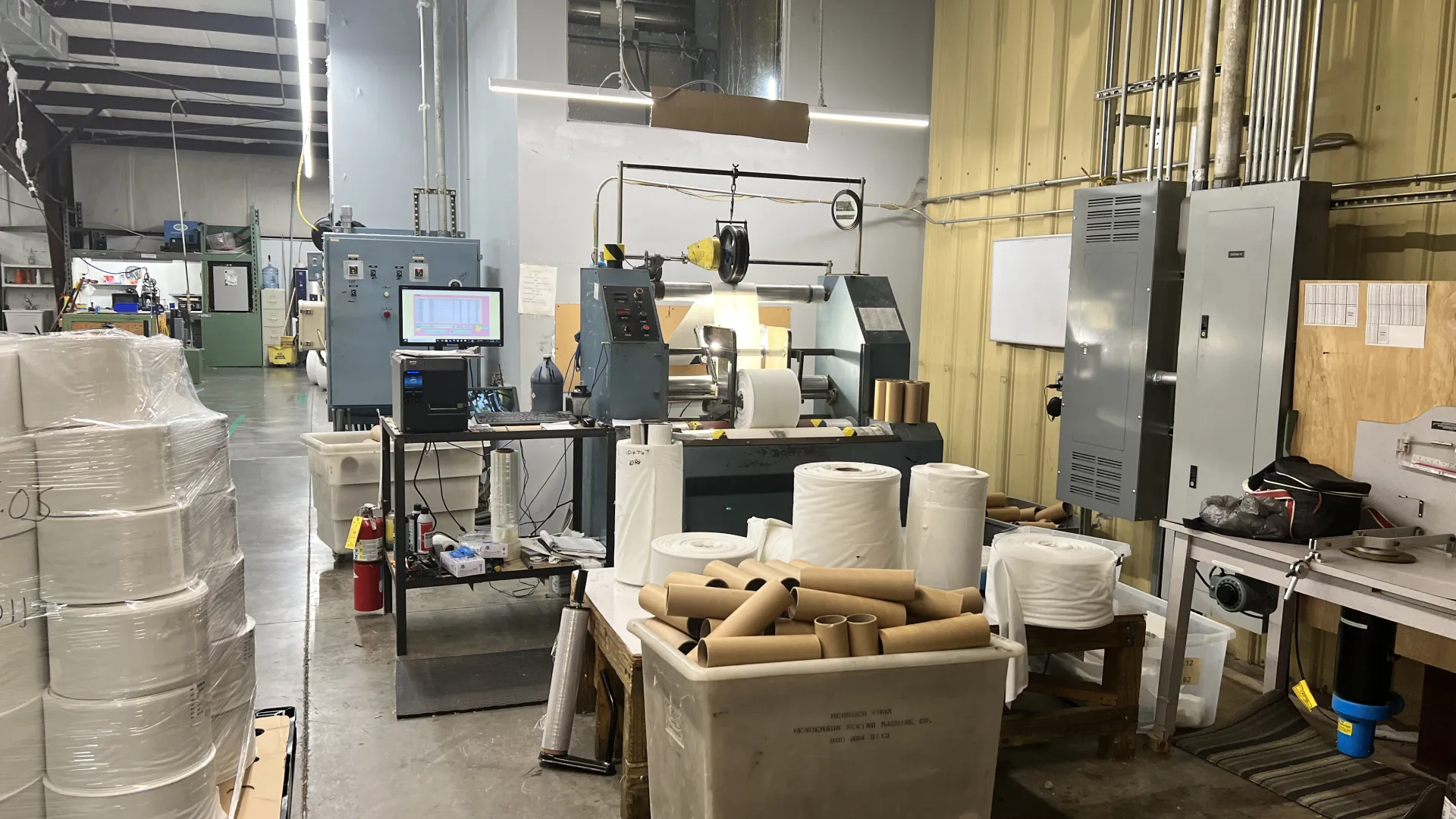Industrial workspace with machinery, rolls of material, cardboard tubes, and control panels in a factory setting