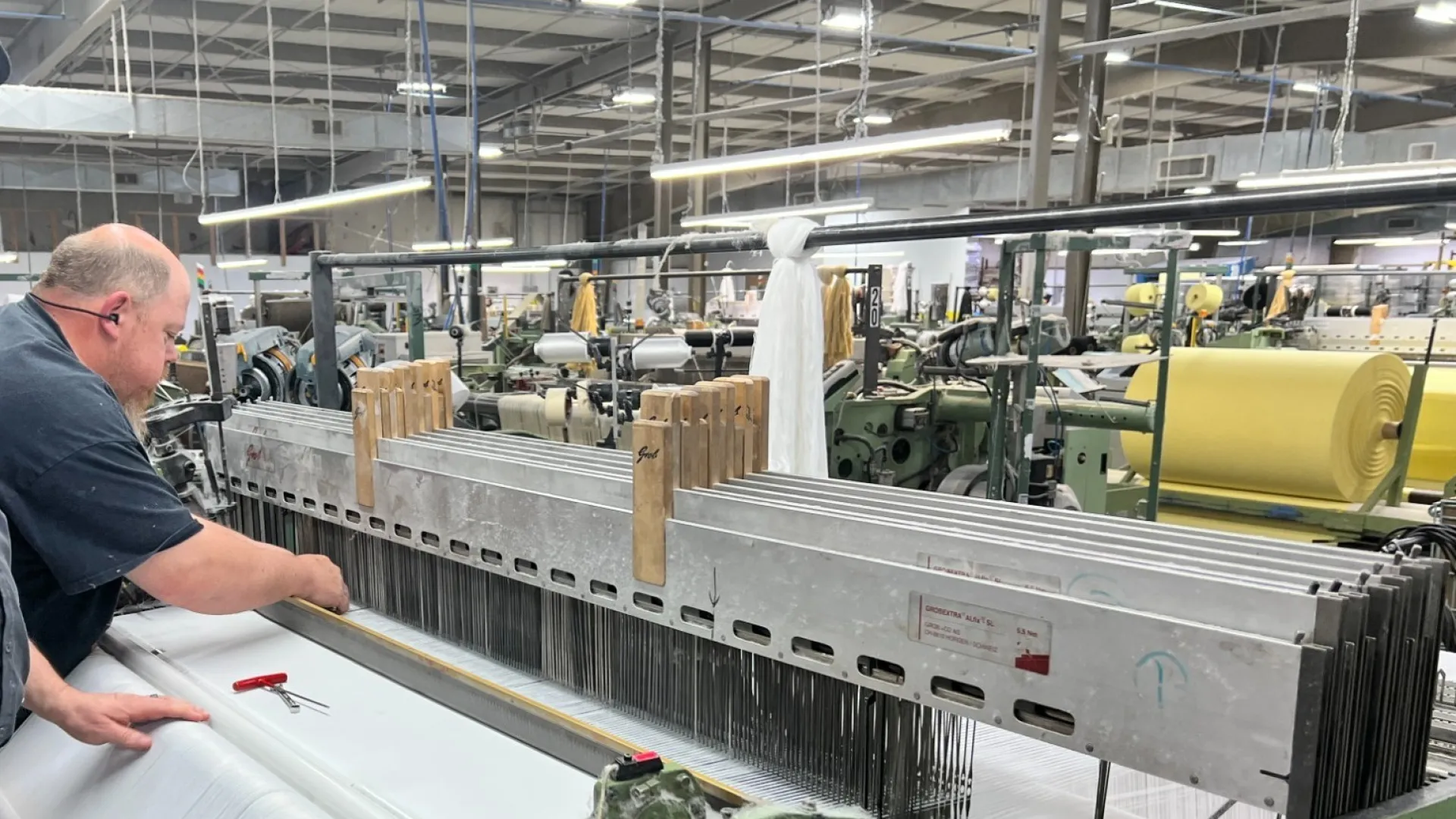 Two workers operate textile machinery in a factory producing white fabric on industrial looms.