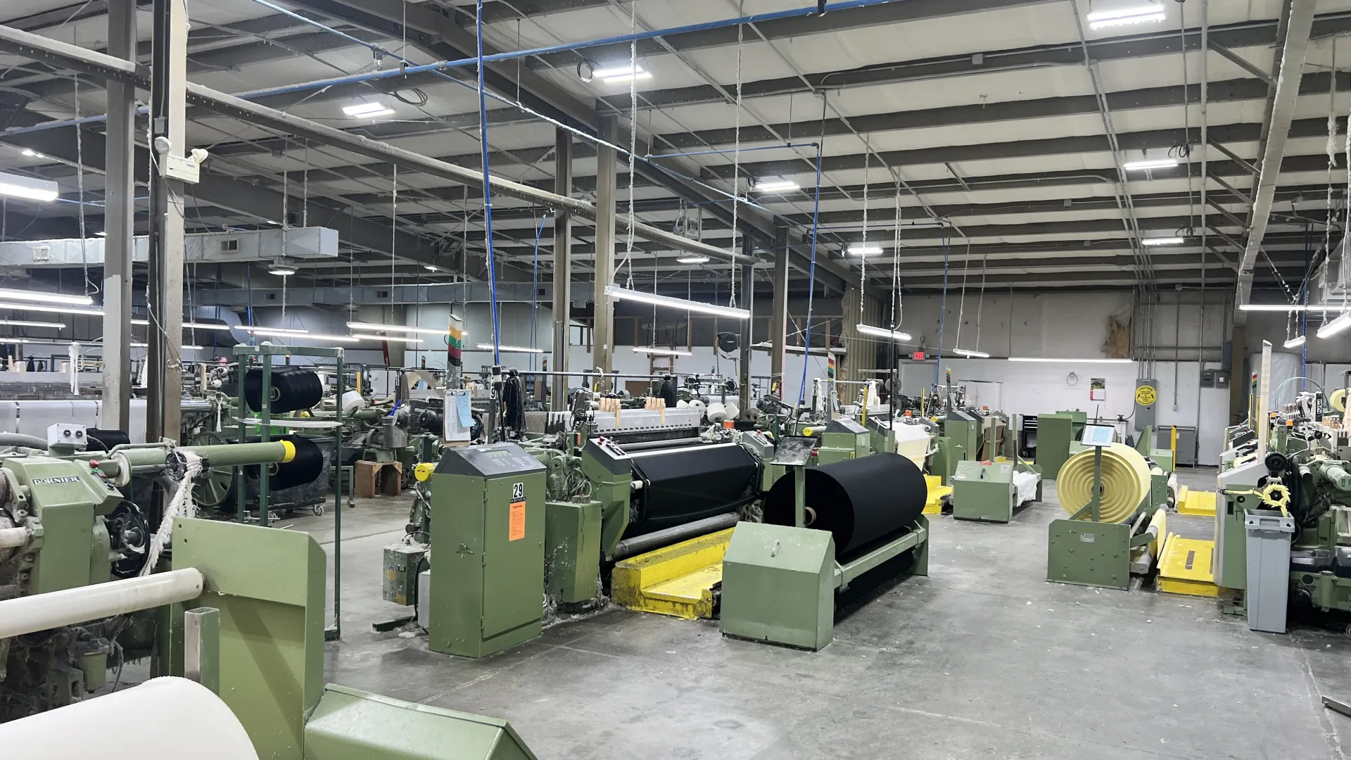 Spacious textile factory floor with rows of industrial weaving machines and fabric rolls under bright lighting