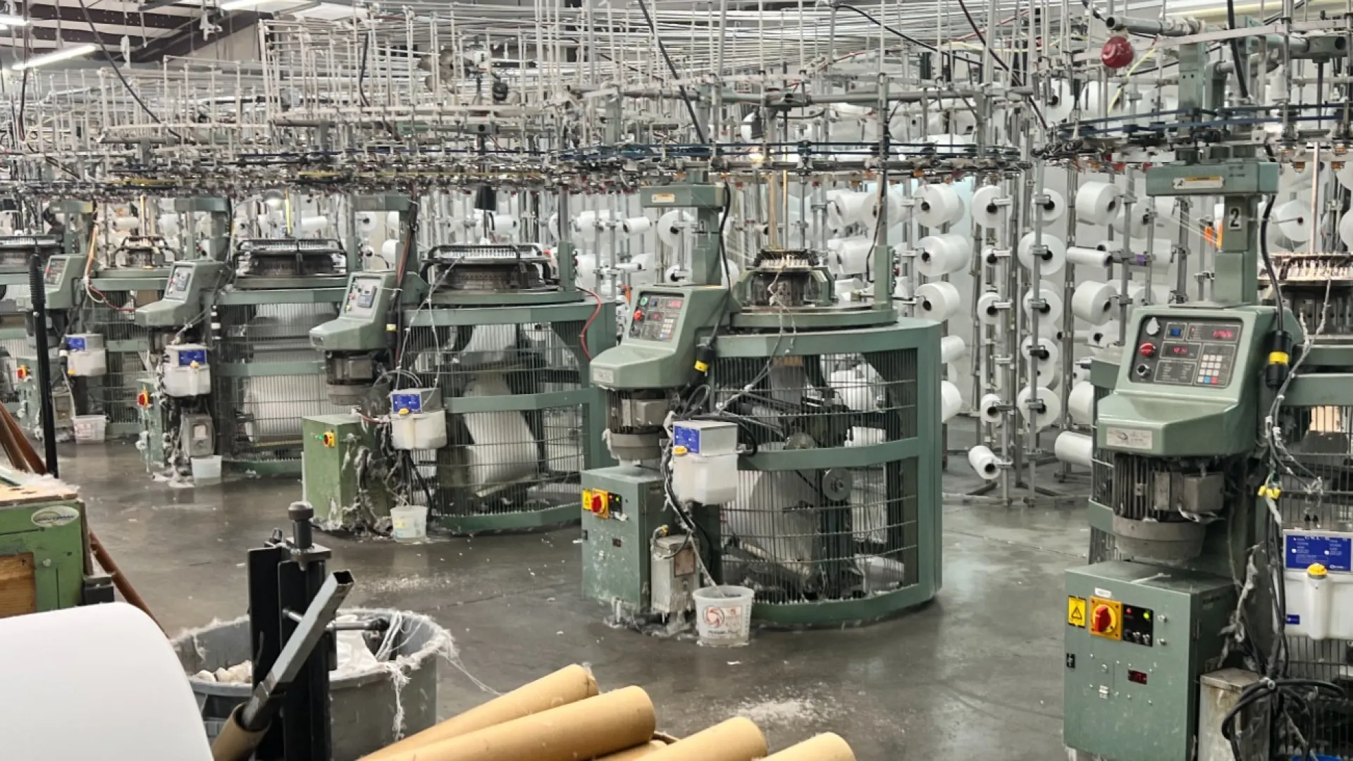 Industrial textile manufacturing facility with multiple circular knitting machines and yarn spools under fluorescent lights