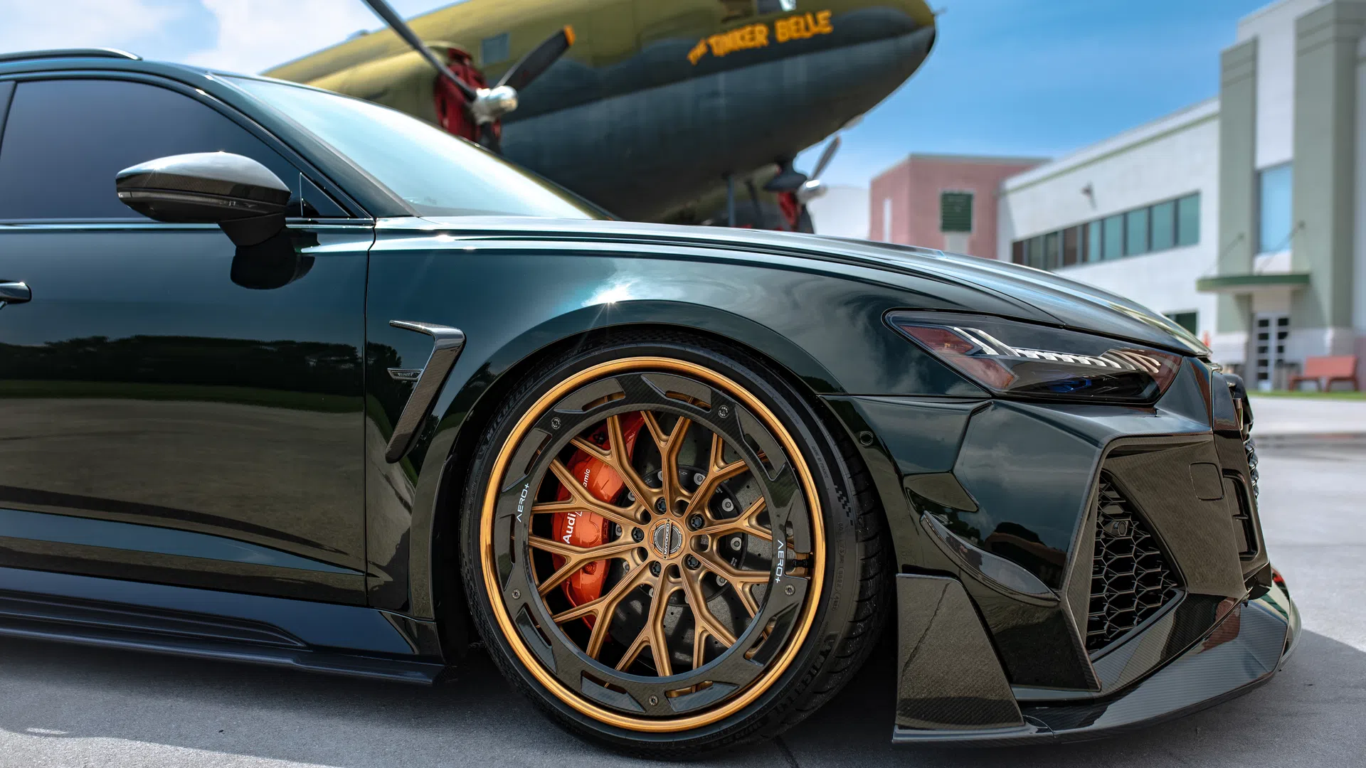 Close-up of a black luxury car with custom gold-rimmed wheels parked near a historic airplane and modern buildings.