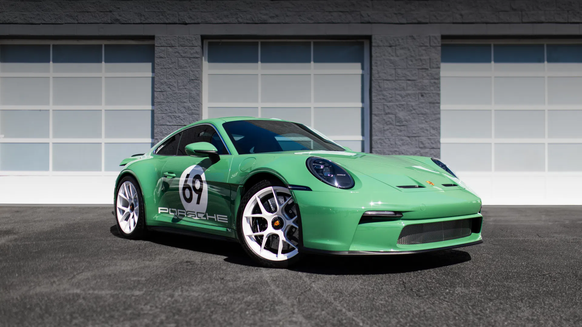 Green Porsche sports car with number 69 decal parked in front of gray building with white garage doors.