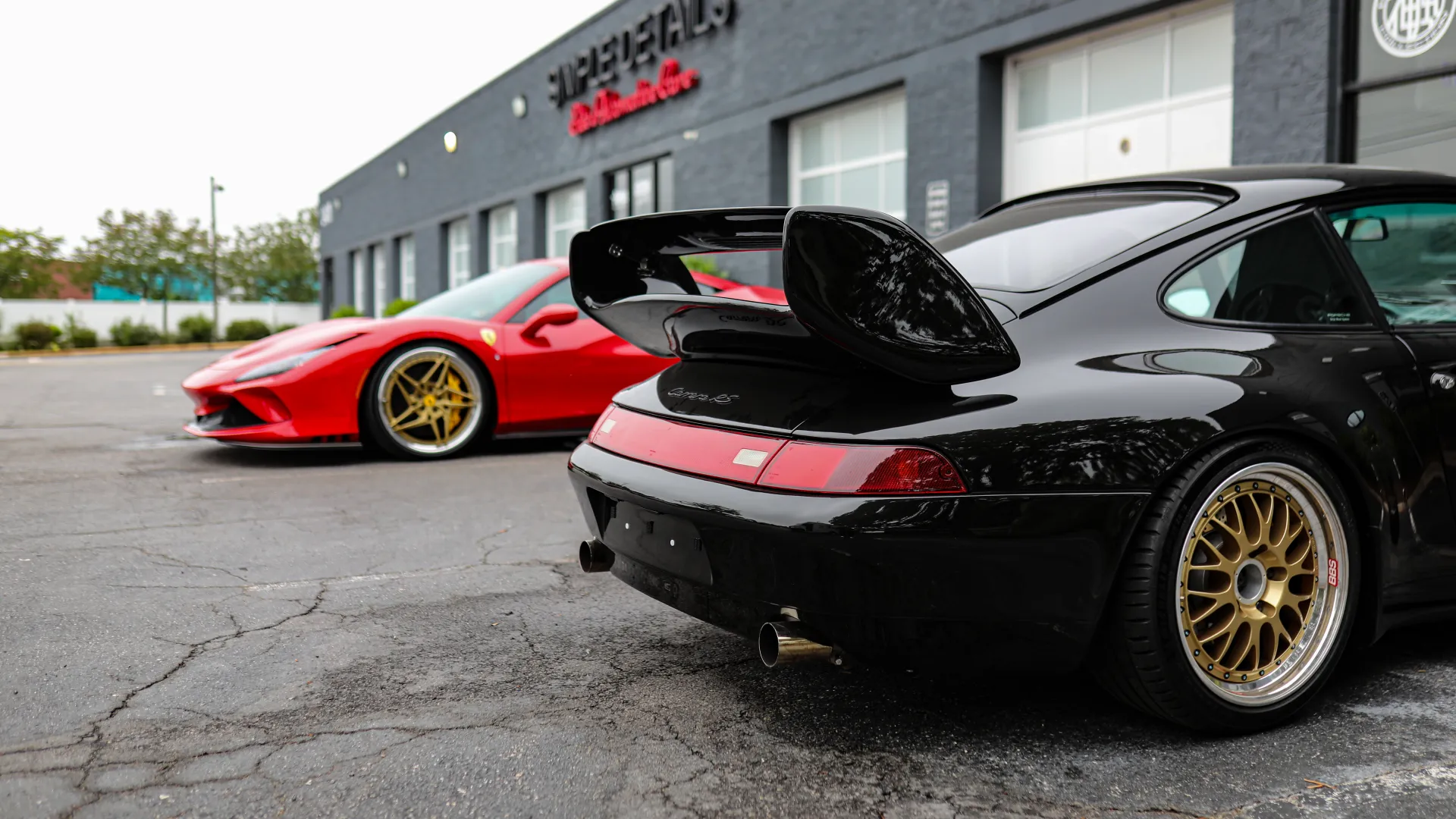 Black Porsche 911 Turbo and red Ferrari sports cars parked outside a tuning shop on cracked asphalt.