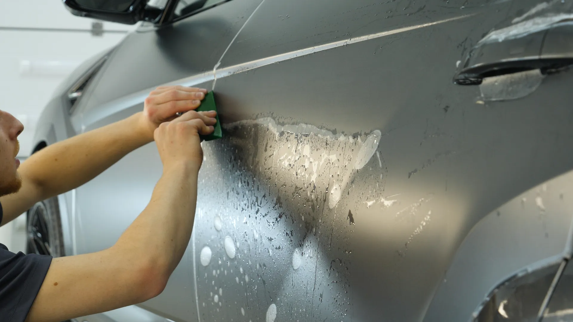 Person applying protective film or vinyl wrap to a sleek silver car door with water droplets