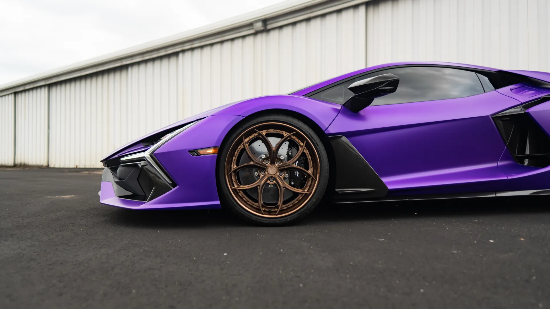 Side view of a sleek purple sports car with bronze alloy wheels parked on black asphalt near a white industrial wall